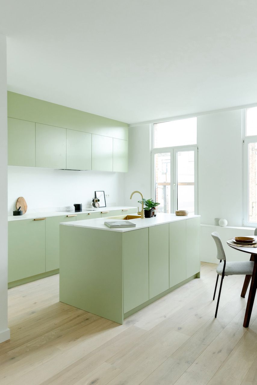 Saliegroene keuken met eiland, wit blad en messing kraan, naast een eettafel bij het raam